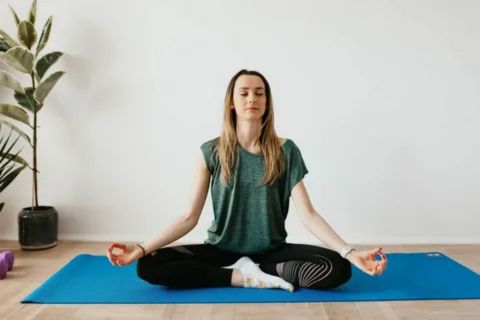 A woman sitting in a lotus position on a blue yoga mat with her eyes closed, practicing mindfulness meditation to manage stress and anxiety.