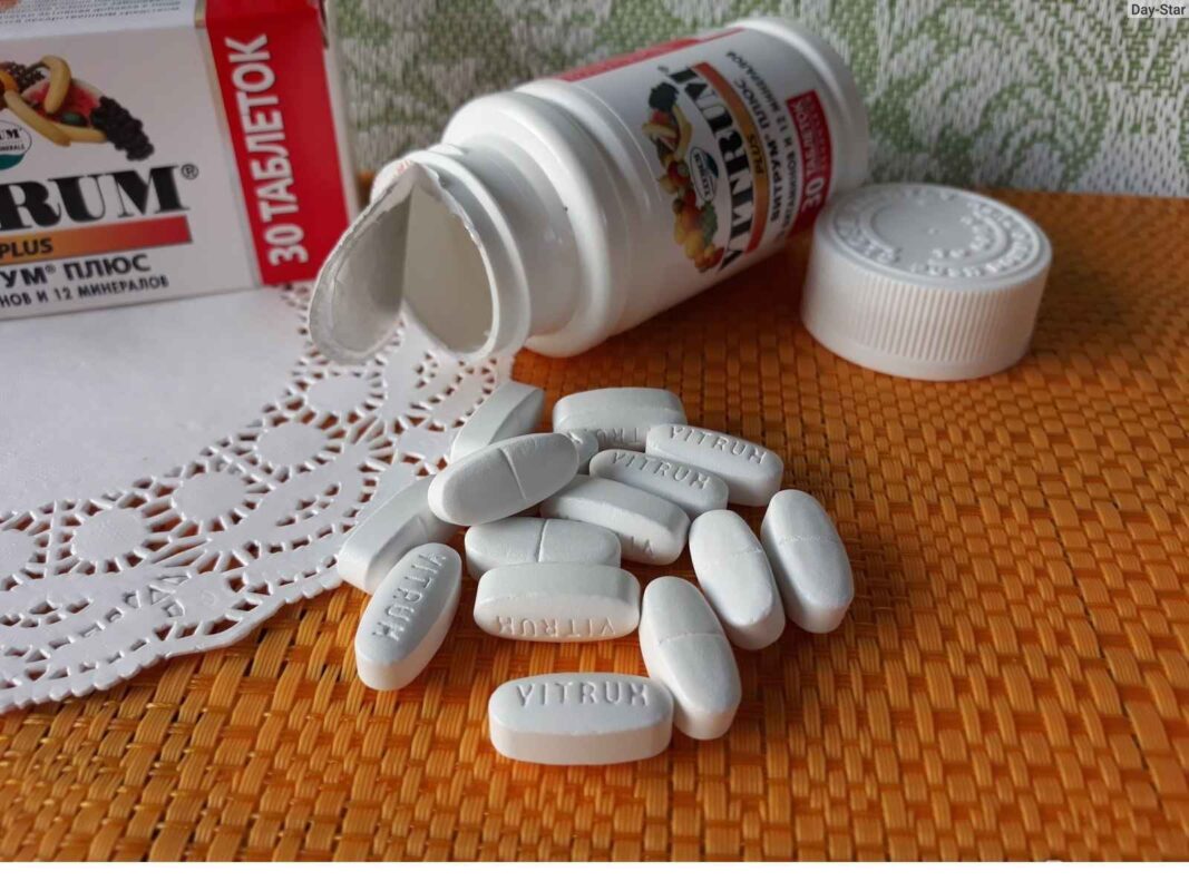 A close-up, low-angle shot of a white plastic bottle of Vitrum Plus vitamins lying on its side. The cap is off, and several white, oval-shaped pills with the word "VITRUM" embossed on them are scattered on a textured orange placemat next to a lacy white coaster. A red and white box is visible in the background.
