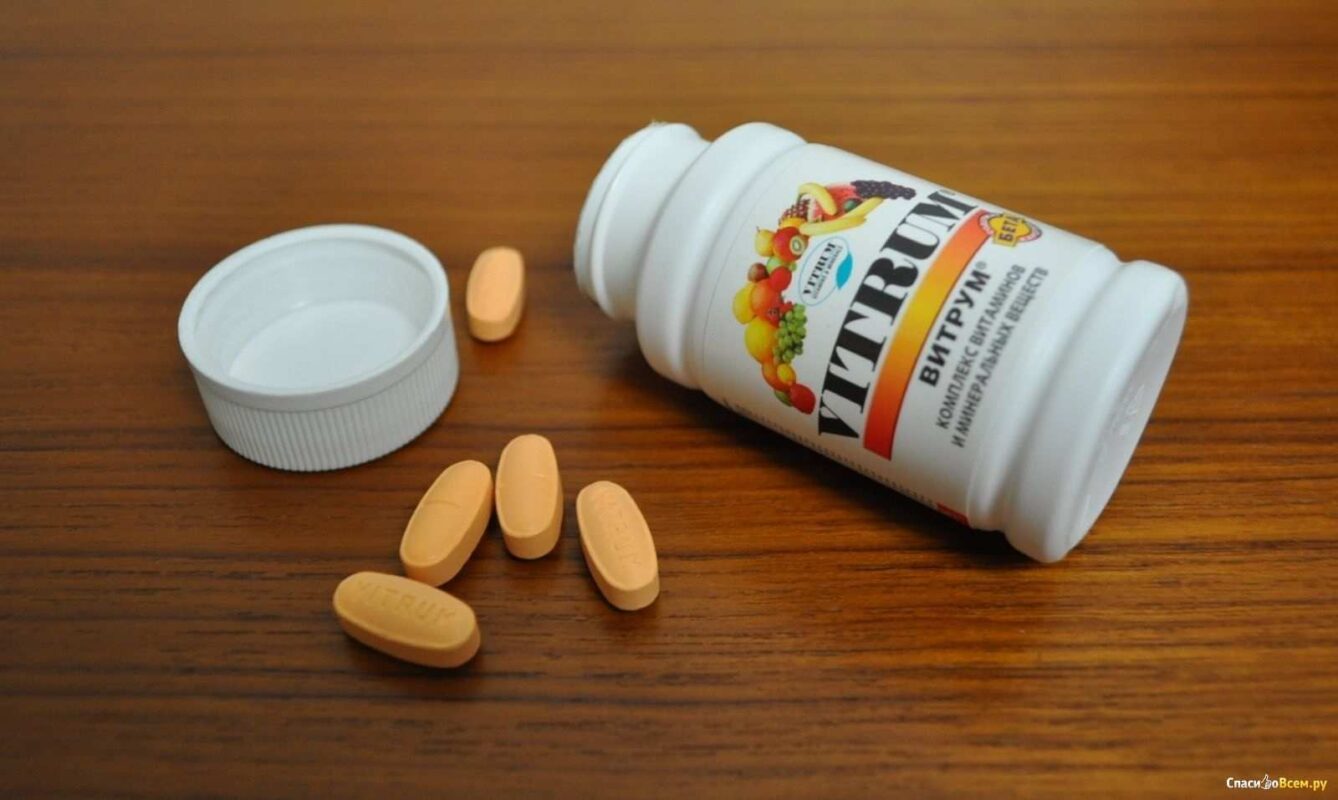 A still life photograph featuring a white bottle of Vitrum Multivitamin Tablets lying on its side on a wooden surface. The label is visible and shows the brand name "VITRUM" along with a picture of various fruits. The cap is removed and lies next to the bottle, with several orange oval-shaped tablets spilled nearby. The focus is sharp on the tablets and the bottle.